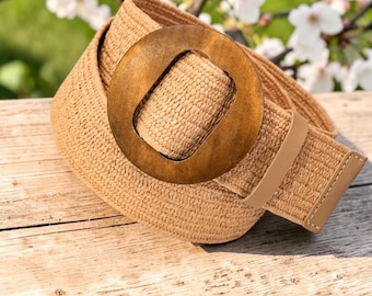 May include: A tan woven belt featuring a large, circular, dark brown wooden buckle. The belt strap is constructed from a textured, braided material and includes a matching tan loop to secure the end of the strap near the buckle.