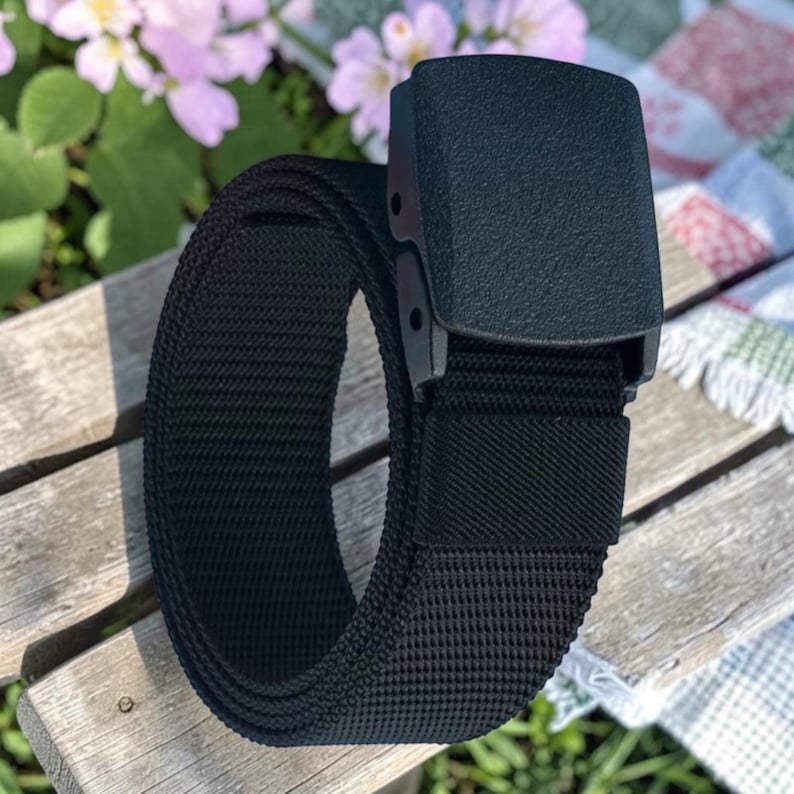 May include: A black nylon webbing belt featuring a rectangular plastic buckle with a textured matte finish. The belt strap is constructed from a durable woven fabric and includes a matching black fabric loop to secure the end of the strap.