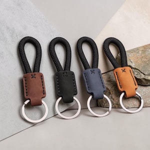 May include: Four keychains featuring a black braided rope loop attached to a rectangular leather tab in brown, black, blue, and tan. Each tab is stitched with a small cross detail and connected to a circular silver-toned metal split ring.