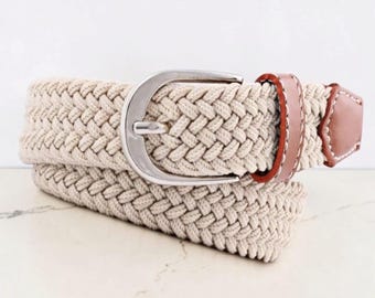May include: A beige braided belt featuring a silver-toned metal buckle and brown leather-like accents with white stitching. The belt is constructed from a woven fabric material and includes a matching brown leather-like tip at the end.