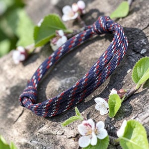 Può includere: Un braccialetto a tema nautico realizzato con una spessa corda intrecciata. La corda è blu navy con un motivo a macchie rosse e bianche. Il braccialetto è annodato e poggia su una superficie di legno stagionato, circondato da fiori bianchi e foglie verdi.