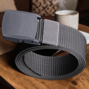 May include: A dark gray tactical belt featuring a textured nylon webbing strap and a matching matte gray plastic buckle. The belt includes a rectangular sliding keeper loop that secures the excess strap, showcasing a durable and functional design.