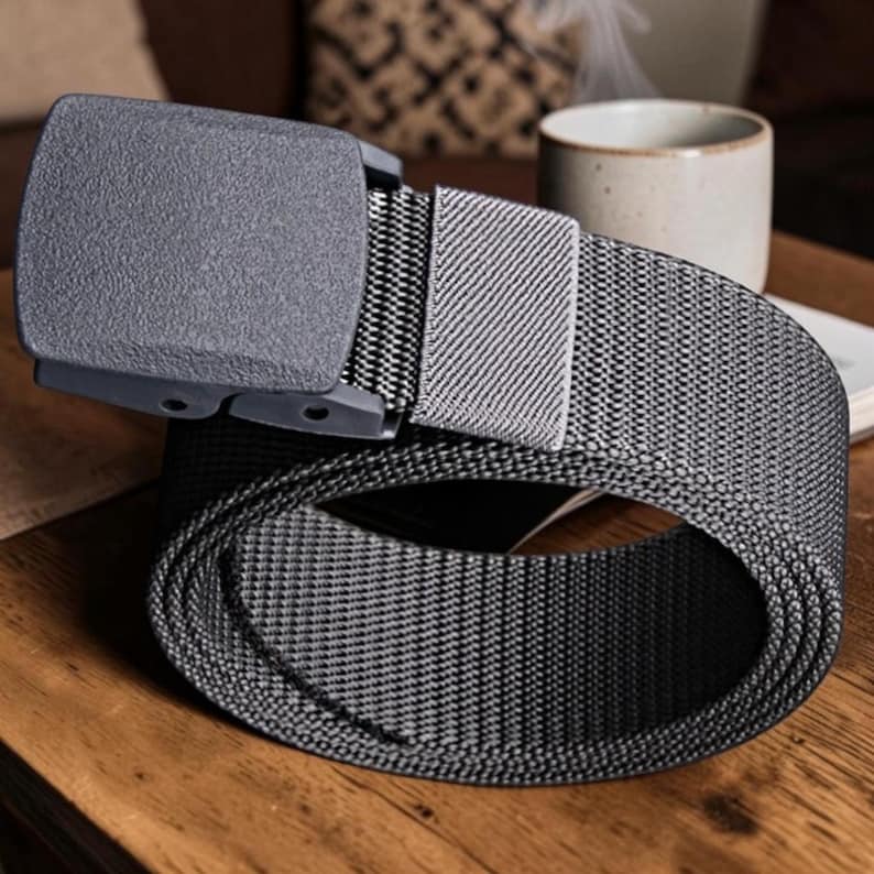May include: A dark gray tactical belt featuring a textured nylon webbing strap and a matching matte gray plastic buckle. The belt includes a rectangular sliding keeper loop that secures the excess strap, showcasing a durable and functional design.