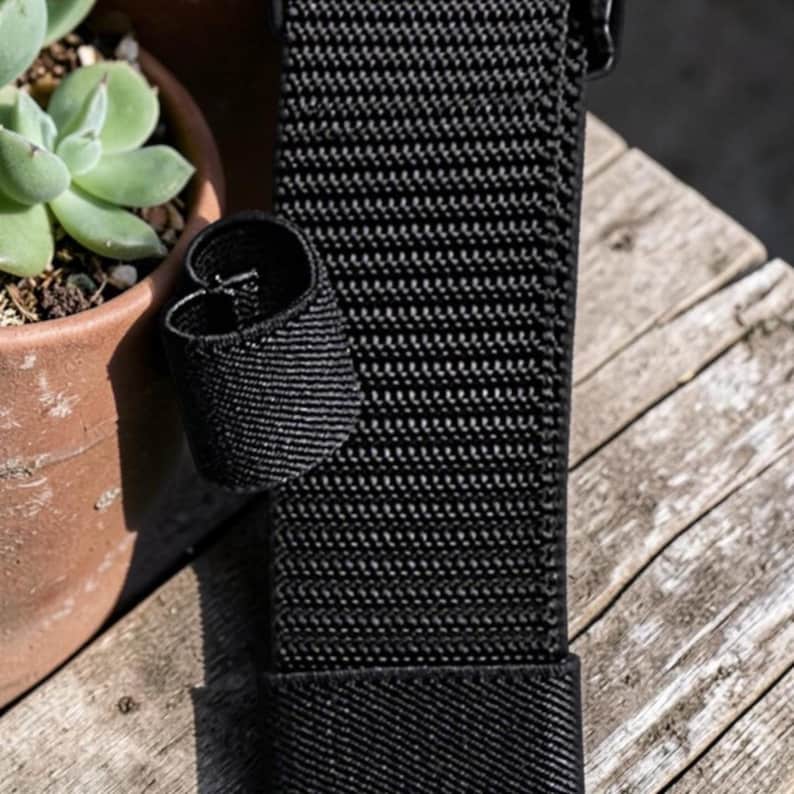 May include: A black nylon webbing strap featuring a textured weave pattern and a small, circular elastic loop attached to the side. The strap is constructed from durable synthetic material with a consistent, tight-knit grid texture throughout its length.