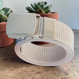 May include: A beige webbed belt featuring a matching rectangular plastic buckle and a single fabric loop. The belt is constructed from a durable woven material with a consistent texture throughout, designed to be adjustable for a secure fit.