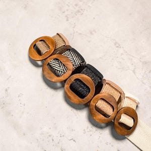 Könnte beinhalten: Eine Sammlung von fünf Gürteln mit runden Holzschließen. Die Gürtel bestehen aus verschiedenen gewebten Stoffen in Beige, Schwarz-Weiß-Fischgrätmuster, Schwarz und Creme, die jeweils mit einer glatten, runden und polierten Holzschließe befestigt sind.