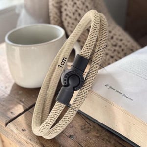 Könnte beinhalten: Ein beiges Seilarmband mit schwarzem Verschluss, ca. 1 cm breit. Das Armband ist geschlungen und liegt auf einer Holzoberfläche neben einer weißen Tasse und einem offenen Buch. Das Seil hat eine gewebte Textur.