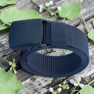May include: A dark navy blue webbed belt featuring a solid, textured, rectangular plastic buckle. The belt strap is constructed from a durable, woven nylon material with a consistent grid pattern throughout the length of the strap.