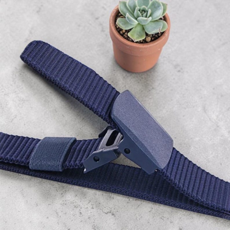 May include: A navy blue woven nylon belt featuring a matching matte plastic buckle with a flip-top closure mechanism. The belt strap has a textured ribbed surface and includes a single fabric loop to secure the excess end of the strap.