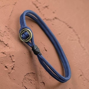 Puede incluir: Una pulsera azul marino con un diseño de cuerda trenzada. La pulsera presenta un cierre cuadrado de color bronce y un detalle de nudo negro. Un accesorio sencillo pero elegante.