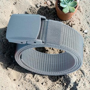 May include: A light gray woven fabric belt featuring a matching matte plastic buckle. The belt is coiled into a circular shape, showcasing the textured weave of the strap and the smooth, solid surface of the rectangular buckle mechanism.