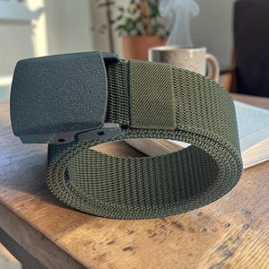 May include: An olive green nylon webbing belt featuring a matte dark green plastic buckle. The belt is coiled into a circular shape, showcasing the textured weave of the fabric strap and the solid, rectangular design of the buckle mechanism.