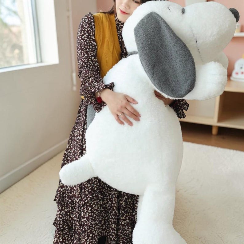 Pu&ograve; includere: Un grande peluche di Snoopy bianco con orecchie grigie. Il peluche &egrave; tenuto da una persona. Il peluche di Snoopy &egrave; un personaggio popolare del fumetto Peanuts.