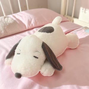 Pu&ograve; includere: Un cuscino a forma di cane bianco e morbido con orecchie grigie e una macchia grigia sulla schiena. Il cane ha gli occhi chiusi e un piccolo naso grigio. &Egrave; appoggiato su un copriletto rosa.