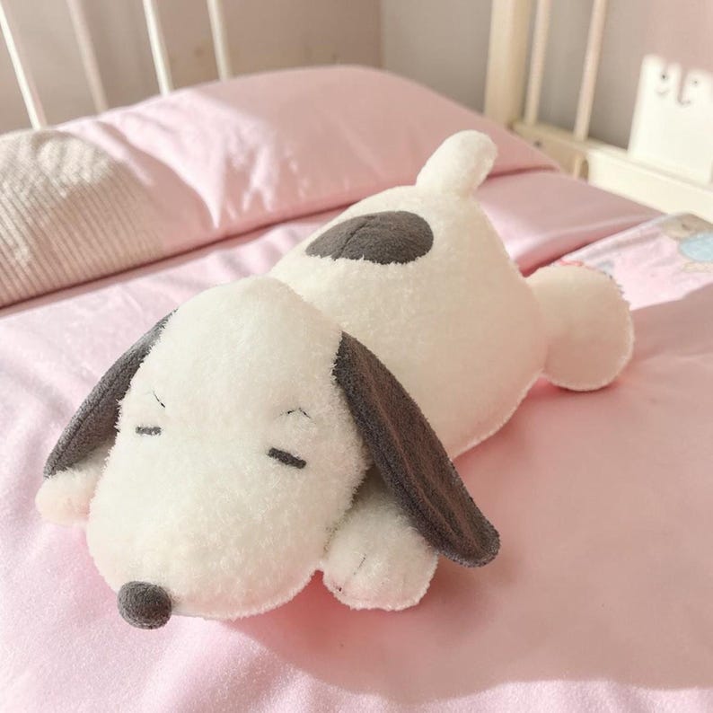 Pu&ograve; includere: Un cuscino a forma di cane bianco e morbido con orecchie grigie e una macchia grigia sulla schiena. Il cane ha gli occhi chiusi e un piccolo naso grigio. &Egrave; appoggiato su un copriletto rosa.