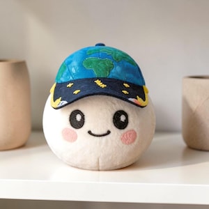 May include: A plush toy shaped like a smiling, round character wearing a blue baseball cap with a map of the world. The toy has a cream-colored body, pink cheeks, and black eyes. The cap has yellow accents.