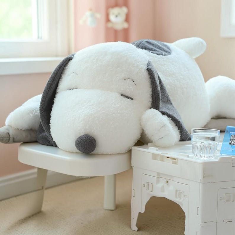 Pu&ograve; includere: Un grande peluche di Snoopy, il cane, in posizione di riposo. Il giocattolo &egrave; principalmente bianco con dettagli grigi sulle orecchie e un naso grigio. &Egrave; appoggiato su un tavolino bianco, suggerendo una cameretta.