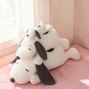 Pu&ograve; includere: Tre peluche bianchi a forma di cane con orecchie e macchie nere sono impilati. I giocattoli sono appoggiati su una superficie rosa, con un piccolo giocattolo sopra due pi&ugrave; grandi. I giocattoli hanno gli occhi chiusi.