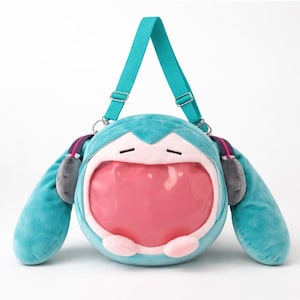 Może przedstawiać: Turkusowa pluszowa torba z dużym otworem w kształcie ust. Torba ma przezroczysty przód, różowe wnętrze i szare akcenty w kształcie uszu. Posiada turkusowy pasek na ramię i jest zaprojektowana tak, aby przypominać postać z kreskówki.