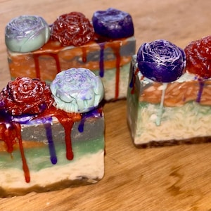 May include: Three rectangular bars of soap featuring layered horizontal bands of white, green, and orange. Each bar is topped with three sculpted flower shapes in purple, red, and light blue, with colorful soap drips running down the sides.