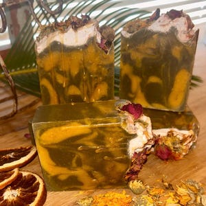 Puede incluir: Tres pastillas de jabón rectangulares hechas a mano con un patrón de remolinos en verde oliva y amarillo dorado. La parte superior de los jabones está decorada con pétalos de flores secas, incluyendo capullos de rosa y caléndulas.
