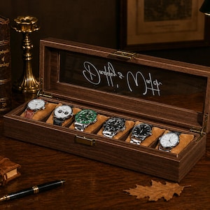 Pode incluir: Uma caixa de madeira escura para relógios com tampa de vidro, contendo seis relógios de pulso com diferentes pulseiras e mostradores. A caixa está sobre uma superfície de madeira, com livros e uma lâmpada no fundo. A tampa de vidro tem escrita cursiva branca.