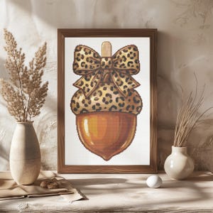 May include: A framed cross-stitch pattern featuring a large acorn with a smooth orange base and a textured cap decorated with a brown and tan leopard print pattern, topped with a matching leopard print bow and a small vertical wooden stem.