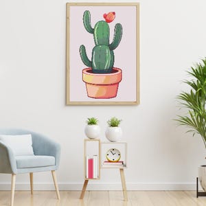 May include: A framed print of a pixelated cactus in a terracotta pot. The cactus is green with a red flower. The artwork is displayed on a light pink background. The room also features a blue chair, a side table with plants, and a floor plant.