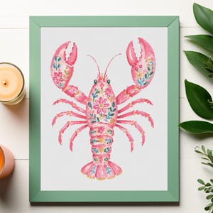 May include: A framed watercolor print of a pink lobster decorated with floral patterns. The artwork is in a mint green frame, with two lit candles and greenery on a white wooden surface.