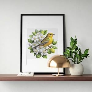 May include: Framed watercolor print of a yellow bird perched on a flowering branch. The bird is surrounded by white blossoms and green leaves. The artwork is displayed in a black frame, resting on a brown shelf with a gold lamp and a potted plant.