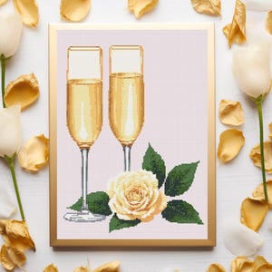 Könnte beinhalten: Kreuzstich-Kunstwerk mit zwei Champagnergläsern, gefüllt mit goldener Flüssigkeit, einer cremefarbenen Rose und grünen Blättern. Das Kunstwerk ist goldgerahmt und vor einem hellrosa Hintergrund platziert. Weiße und goldene Blütenblätter und weiße Blumen umgeben das gerahmte Kunstwerk.