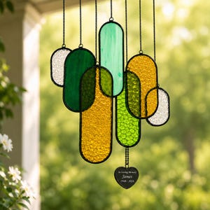 Puede incluir: Un carillón de viento de vidrieras con múltiples paneles ovalados de vidrio texturizado verde, amarillo y transparente, suspendidos por cadenas negras. Un pequeño dije negro en forma de corazón cuelga en la parte inferior con el texto In Loving Memory James 1948-2024.