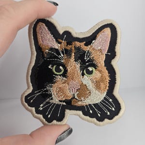 May include: An embroidered patch featuring the head of a calico cat with black, orange, and white fur. The cat has green eyes, a pink nose, and white whiskers. The patch has a black background and a thick, light beige stitched border.