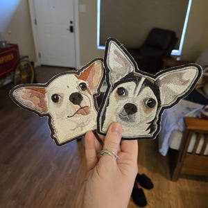 May include: Two embroidered patches featuring dog faces. One patch depicts a light-colored dog with brown ears and markings. The other patch shows a black and white dog. Both patches have a brown border.