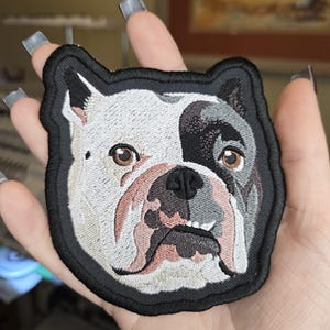 May include: An embroidered patch featuring the head of a bulldog with a white and grey coat. The dog has brown eyes, a black nose, and a black border surrounding the entire shape of the head. The embroidery shows detailed facial features.