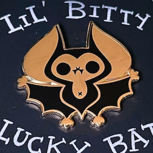 May include: A black and gold enamel pin featuring a cartoon bat with the text "Lil' Bitty Lucky Bat" and "By Arica Tuesday" on a black background.