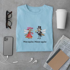 May include: Light blue t-shirt featuring a cartoon couple of mosquitoes, "Miss-squito" in a pink hat and red dress, and "Mister-squito" in a tuxedo. The text "Miss-squito / Mister-squito" is below the graphic. Other items include glasses, a phone, and a coffee cup.