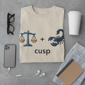 Op de afbeelding: Beige T-shirt met een Weegschaal en Schorpioen cusp ontwerp. De afbeelding toont een weegschaal, een schorpioen en het woord "cusp". Het shirt ligt plat met een telefoon, bril, koptelefoon, koffiebeker en een krant.