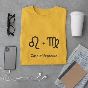 Peut inclure: T-shirt jaune moutarde avec le texte "Leo + Vierge Cusp of Exposure" en noir. Le t-shirt est entouré d'un smartphone, de lunettes, d'une tasse à café, d'écouteurs, d'un carnet et d'un journal.