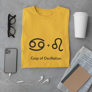 May include: Mustard yellow t-shirt with a black graphic of two zodiac symbols and the text "Cusp of Oscillation." The shirt is surrounded by a smartphone, eyeglasses, a coffee cup, and a newspaper.