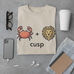 May include: Beige t-shirt with a graphic of a crab, a plus sign, and a lion, with the word "cusp" underneath. The shirt is surrounded by a smartphone, glasses, headphones, a coffee cup, and a newspaper.