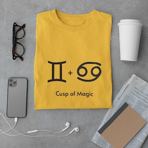 May include: Mustard yellow t-shirt featuring a black Gemini and Cancer zodiac symbol graphic and the text "Cusp of Magic." The image also includes a smartphone, glasses, a coffee cup, and a newspaper.