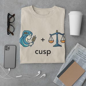 May include: Beige t-shirt with a graphic featuring a woman with blue hair holding wheat, a scale, and the word "cusp". The shirt is surrounded by a smartphone, glasses, headphones, a coffee cup, and a newspaper.
