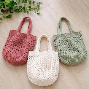 Può includere: Tre borse tote all'uncinetto fatte a mano nei toni rosa antico, crema e verde salvia. Le borse hanno un design a trama aperta e manici robusti. Sono esposte su una superficie di legno chiaro, mettendo in risalto la loro natura artigianale.