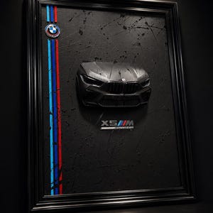 May include: A framed wall art piece featuring a three-dimensional matte black BMW X5 M Competition front end sculpture. The black background has paint splatters, a BMW logo, and vertical light blue, dark blue, and red stripes with X5 M text.