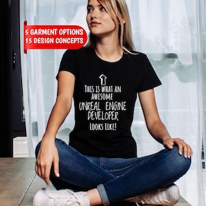 Op de afbeelding: Een zwart t-shirt met korte mouwen, een witte pijl die naar boven wijst en de tekst: This is what an awesome Unreal Engine developer looks like! Een rode banner linksboven toont de tekst: 5 garment options 15 design concepts.