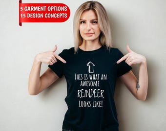 May include: A black short-sleeved t-shirt featuring a white upward-pointing arrow graphic and the text "THIS IS WHAT AN AWESOME REINDEER LOOKS LIKE!" printed on the front. The shirt is paired with dark gray denim jeans.