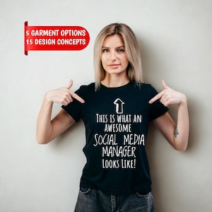 May include: A black short-sleeved t-shirt featuring a white upward-pointing arrow graphic and the text: THIS IS WHAT AN AWESOME SOCIAL MEDIA MANAGER LOOKS LIKE! A red banner in the upper left corner displays the text: 5 GARMENT OPTIONS 15 DESIGN CONCEPTS.
