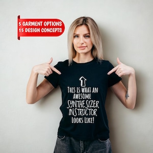 Könnte beinhalten: Ein schwarzes kurzärmeliges T-Shirt mit einer weißen, nach oben zeigenden Pfeilgrafik und dem Text "THIS IS WHAT AN AWESOME SYNTHESIZER INSTRUCTOR LOOKS LIKE!" in einer serifenlosen Schrift auf der Vorderseite des Kleidungsstücks.