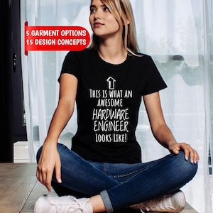 May include: A black short-sleeved t-shirt featuring a white upward-pointing arrow and the text "THIS IS WHAT AN AWESOME HARDWARE ENGINEER LOOKS LIKE!" in a casual font. A red banner in the upper left corner displays "5 GARMENT OPTIONS 15 DESIGN CONCEPTS".